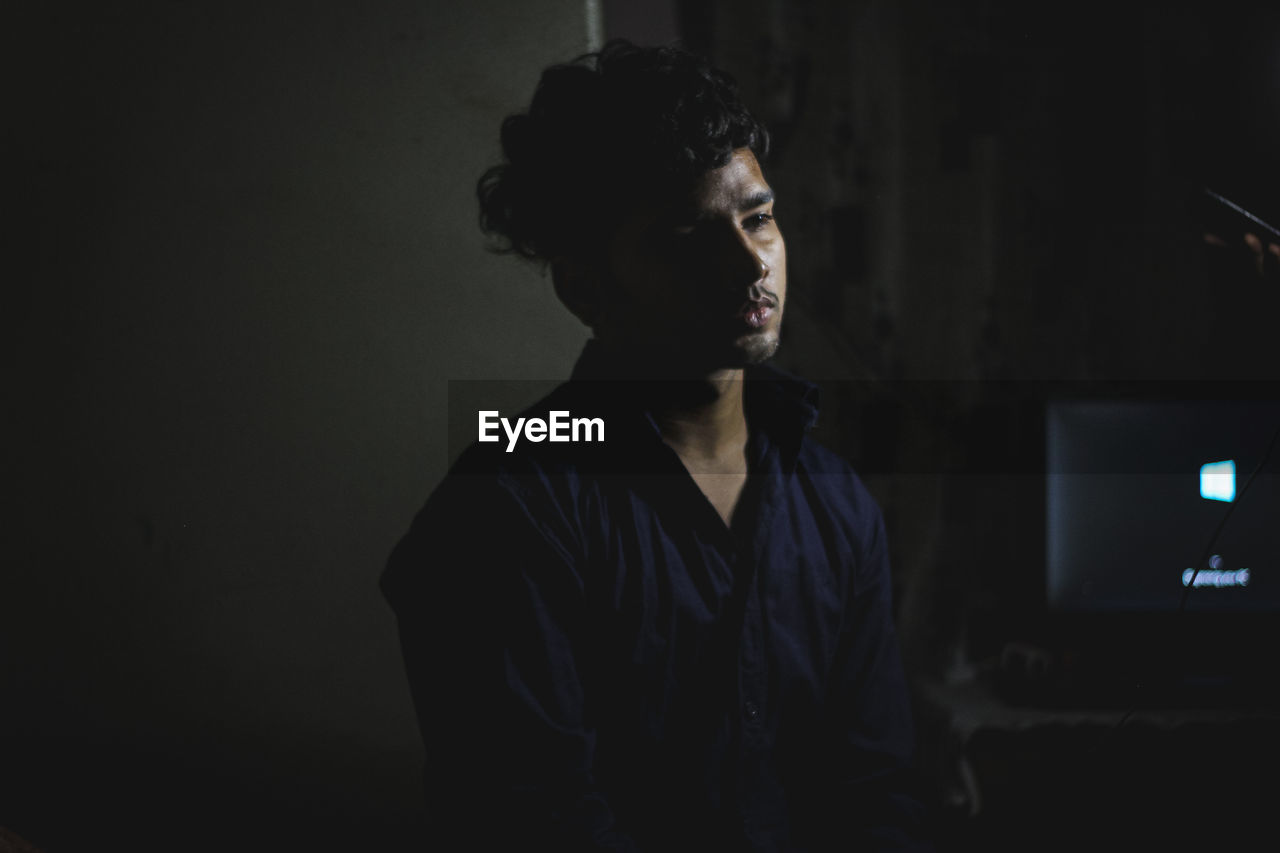 PORTRAIT OF YOUNG MAN LOOKING AWAY WHILE STANDING IN DARK