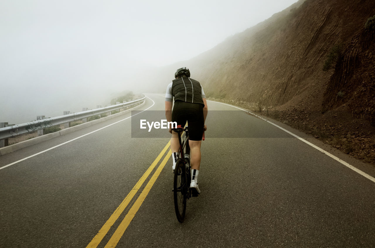 Man cycling on road by mountain during foggy weather