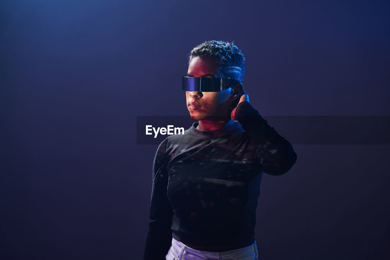 Serious african american female with short hair wearing contemporary vr glasses while standing on purple background in studio with glowing neon lights