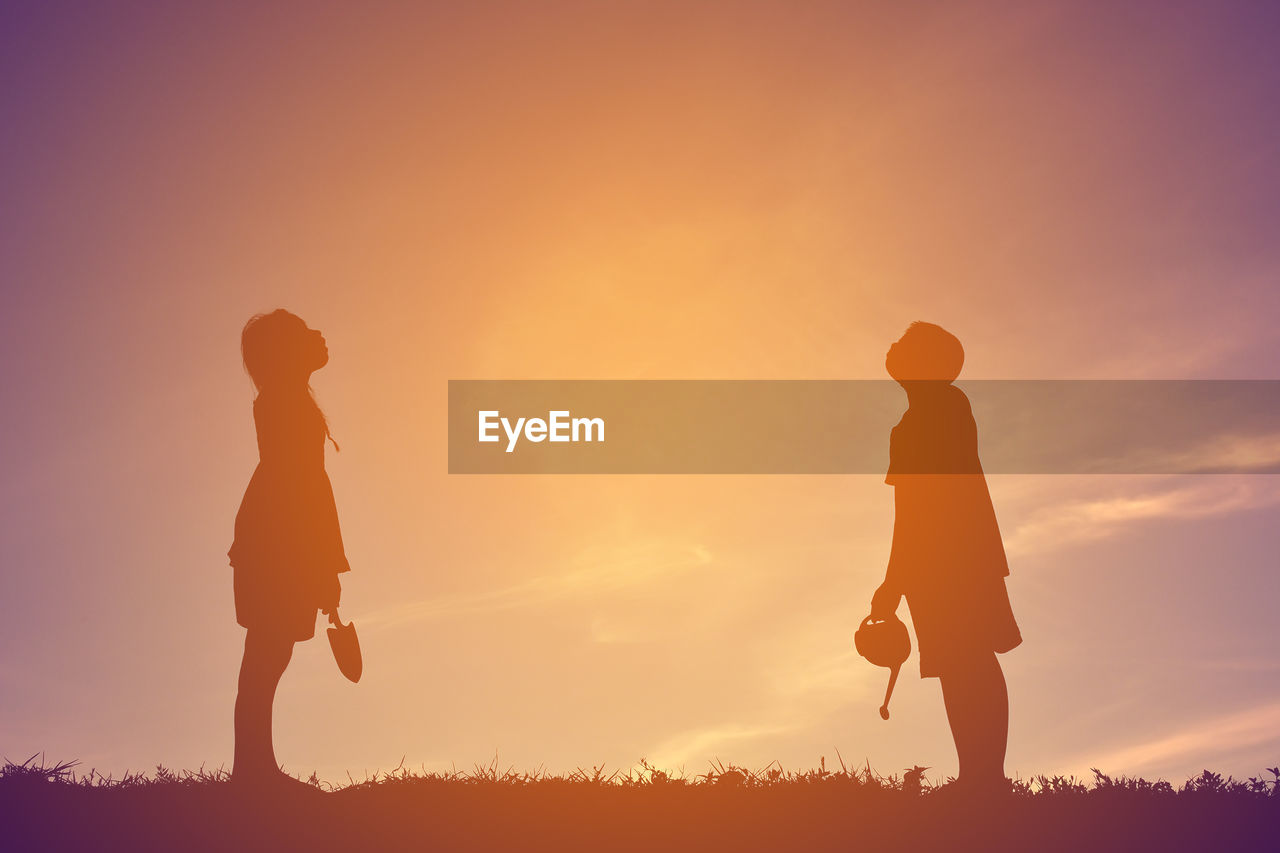 Teenage boy and girl standing with gardening equipment on field against sky during sunset