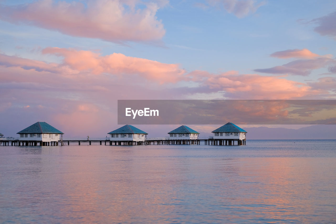 STILT HOUSES BY SEA AGAINST SKY DURING SUNSET