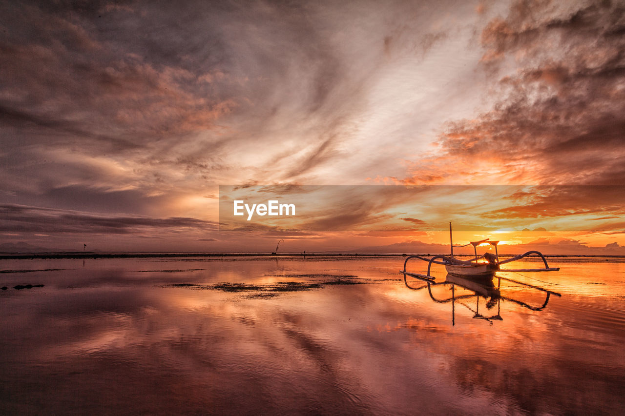 Outrigger boat at beach against cloudy sky during sunset