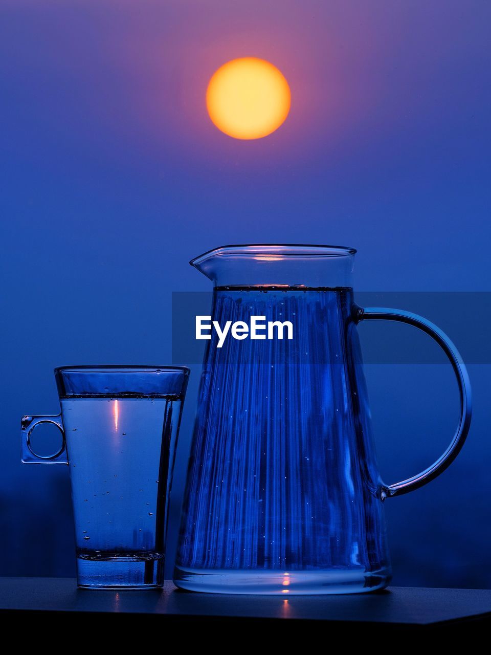 A water jug and drinking glass containing water reflecting distant buildings at sunrise