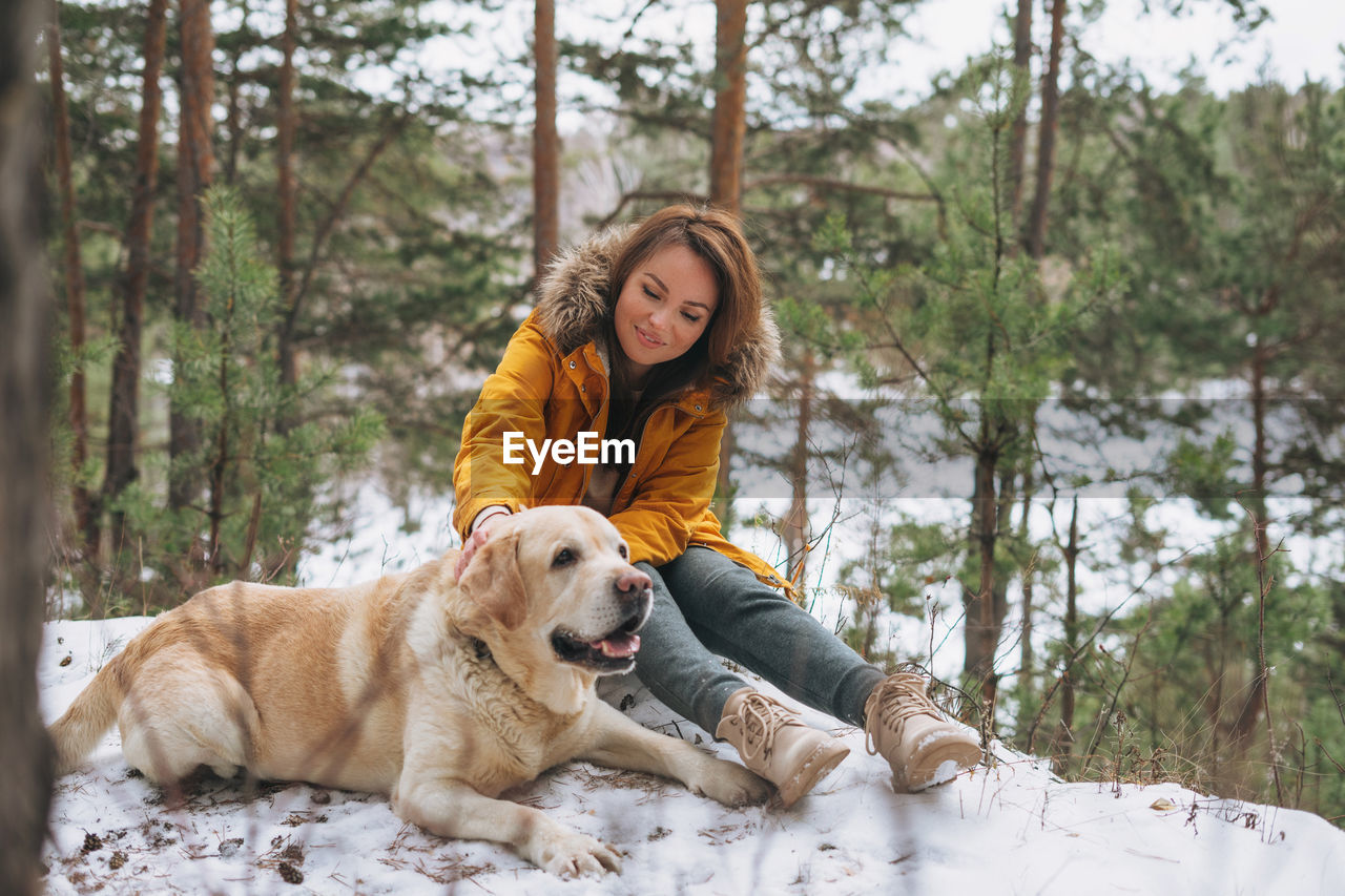 Young smiling woman in yellow jacket with big kind white dog labrador walking in winter forest