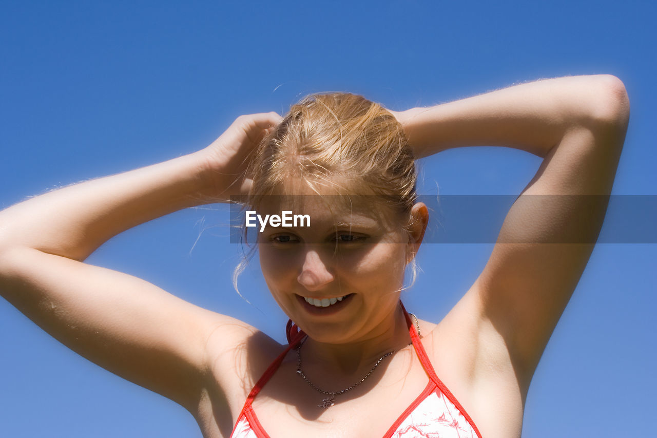 Low angle view of smiling woman with hands behind head against clear blue sky
