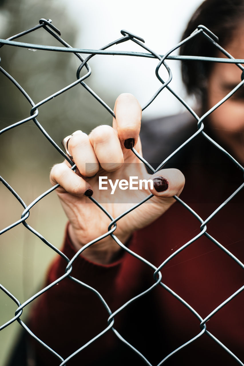 Hand of a woman in red sweater with dark manicured nails pushing against rustical metal fence. 