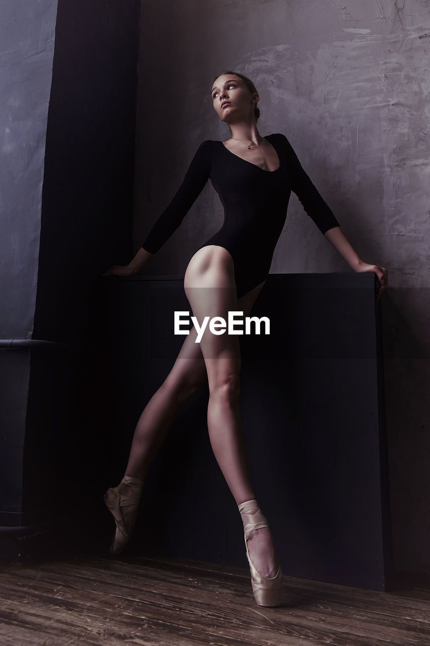 A ballerina in a bodysuit and pointes is standing in the corner of the room looking out the window