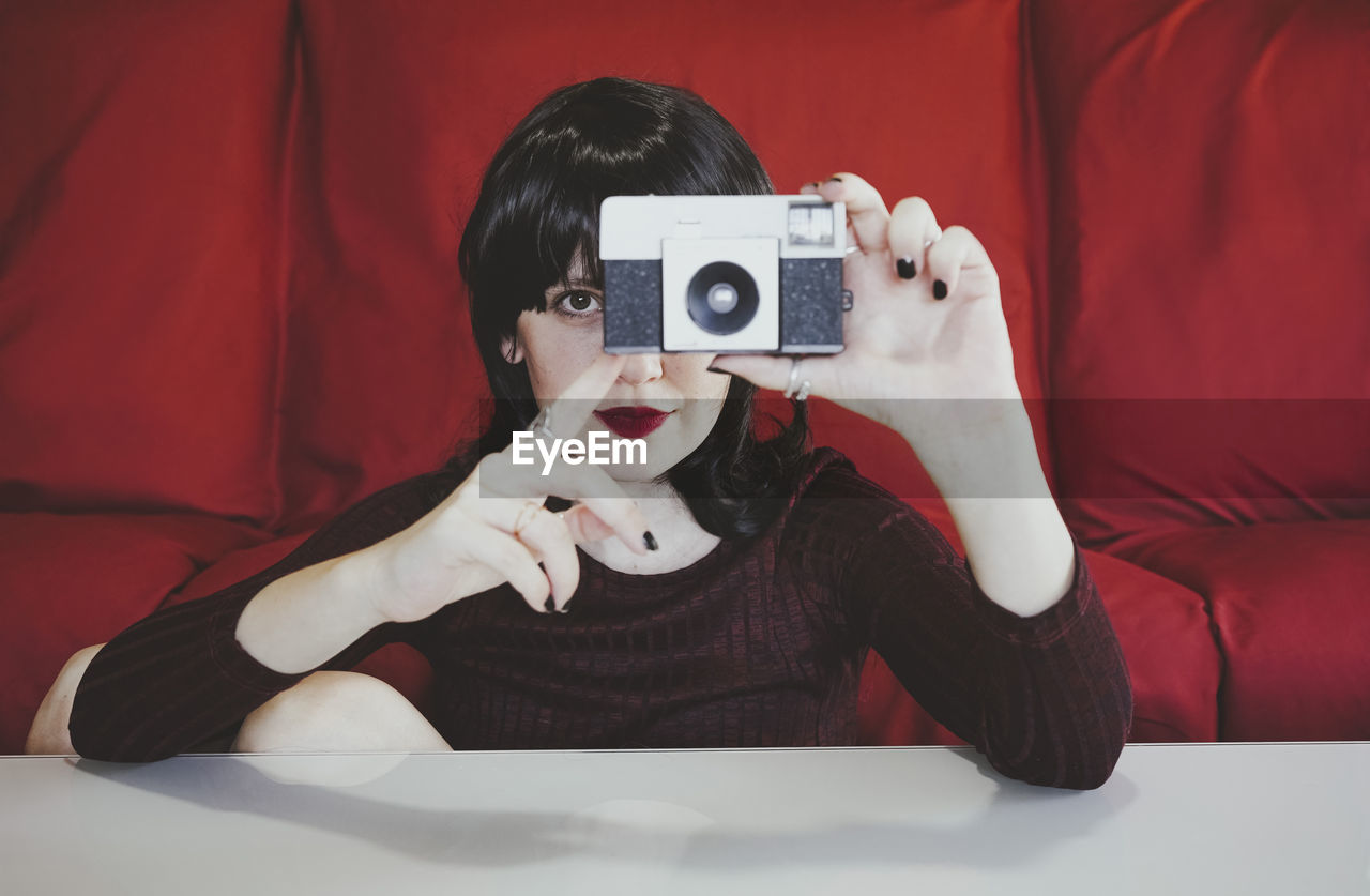 Woman covering face with analog camera in front of red couch