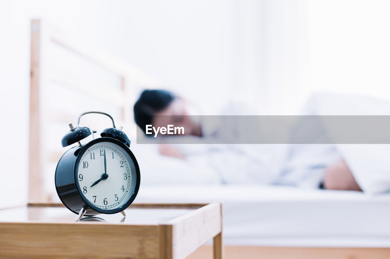 Close-up of alarm clock on side table with woman sleeping in background