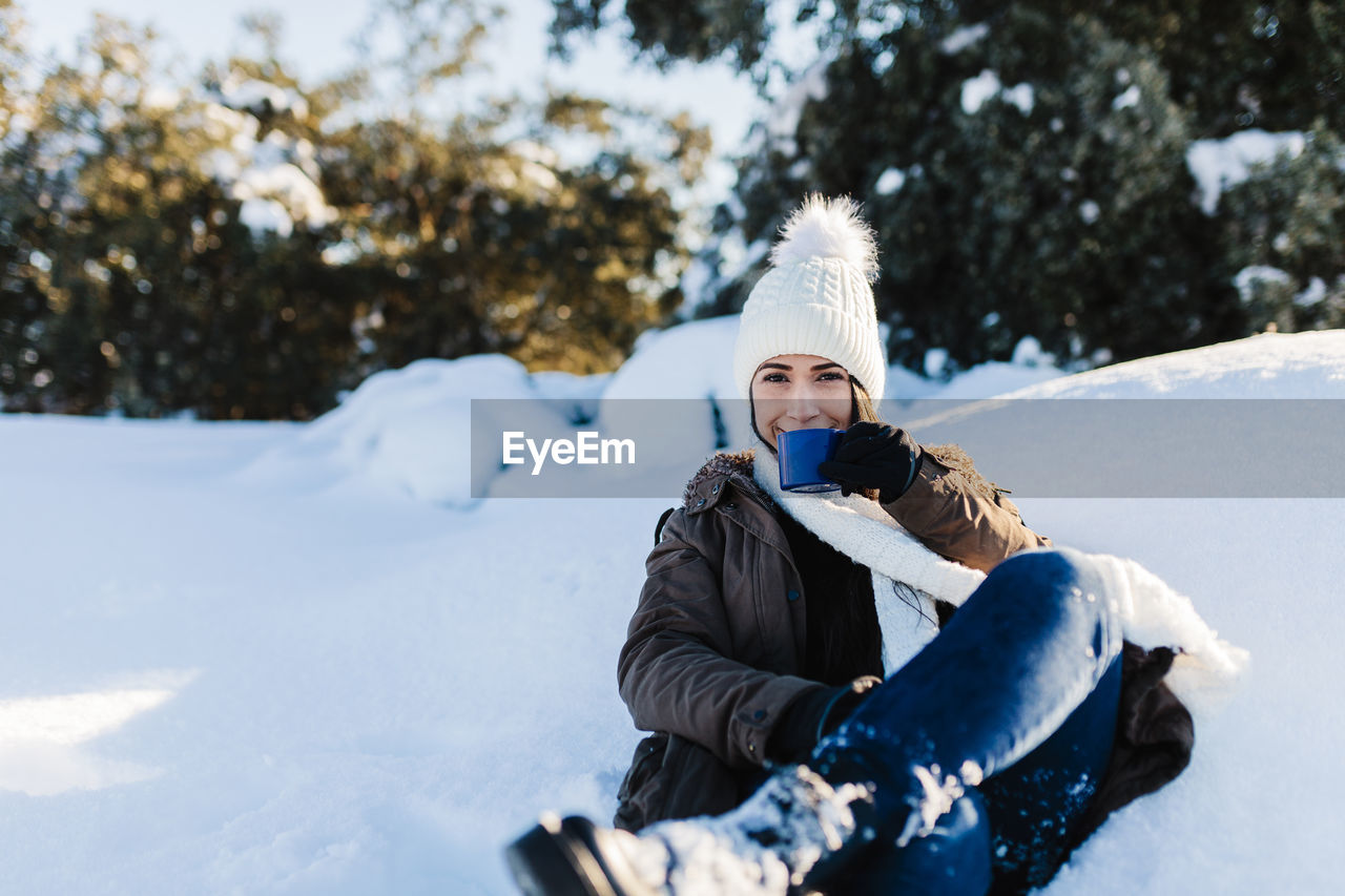 Smiling woman drinking coffee while sitting on snow