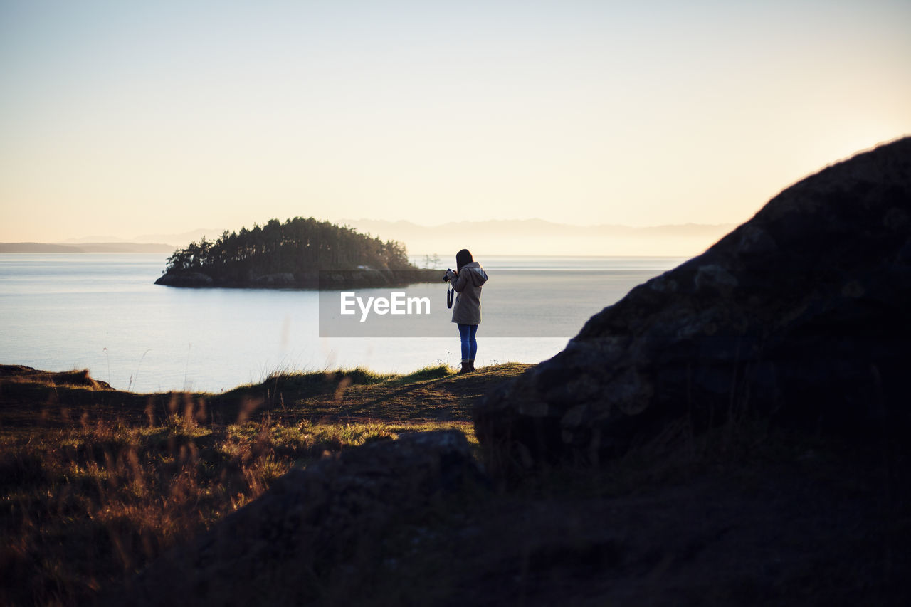 MAN STANDING ON ROCK AT SEA SHORE AGAINST SKY