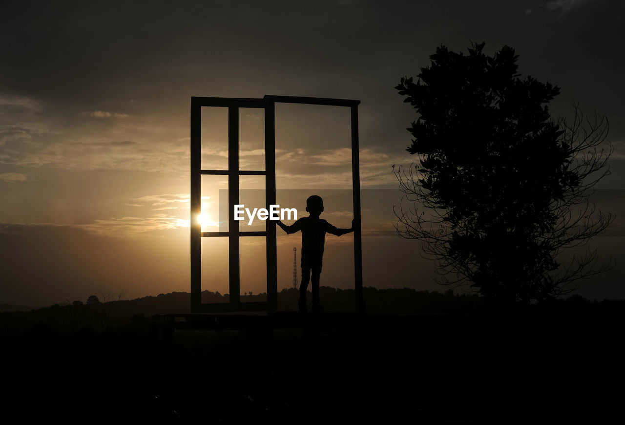 Silhouette boy standing at gate on field against sky during sunset