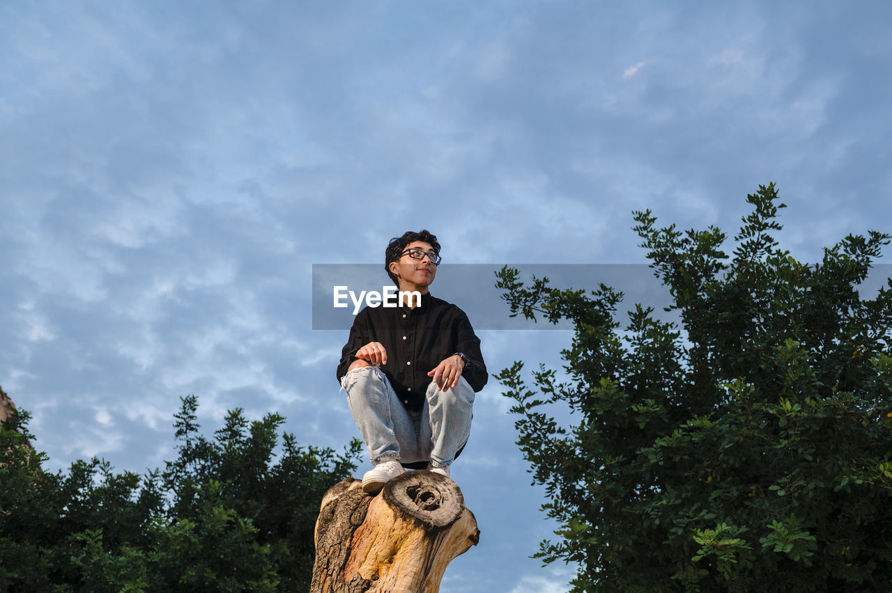 Young transgender man with glasses posing on a log outdoors.