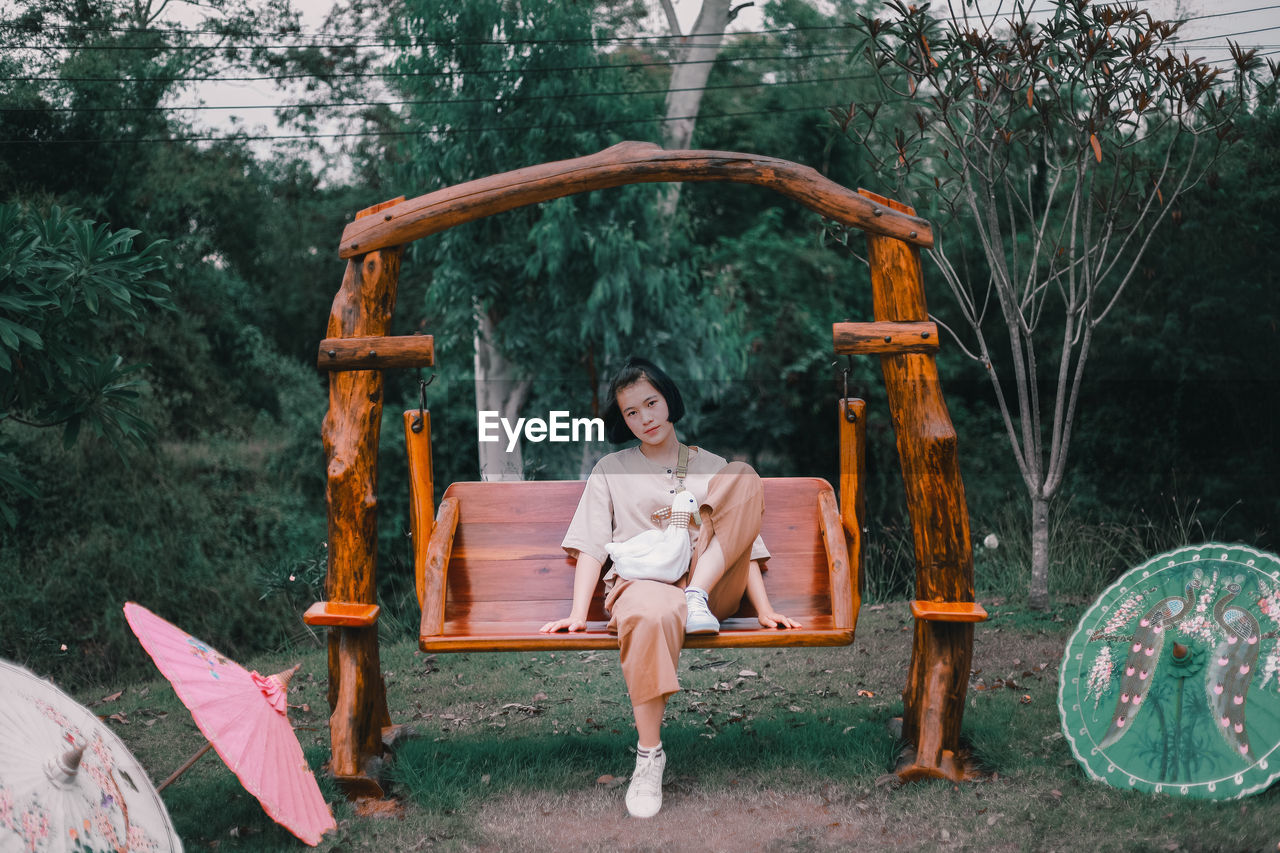 PORTRAIT OF YOUNG WOMAN SITTING AGAINST PLANTS AND TREES