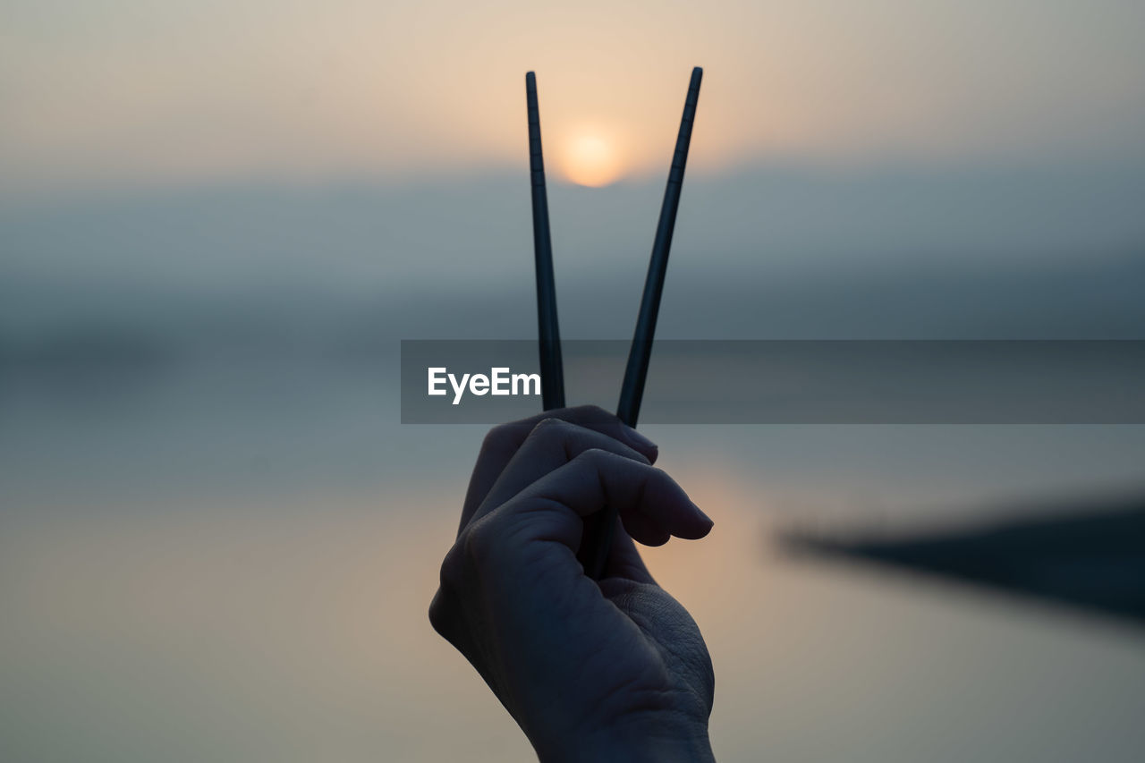Close-up of person holding chopsticks against sky during sunset