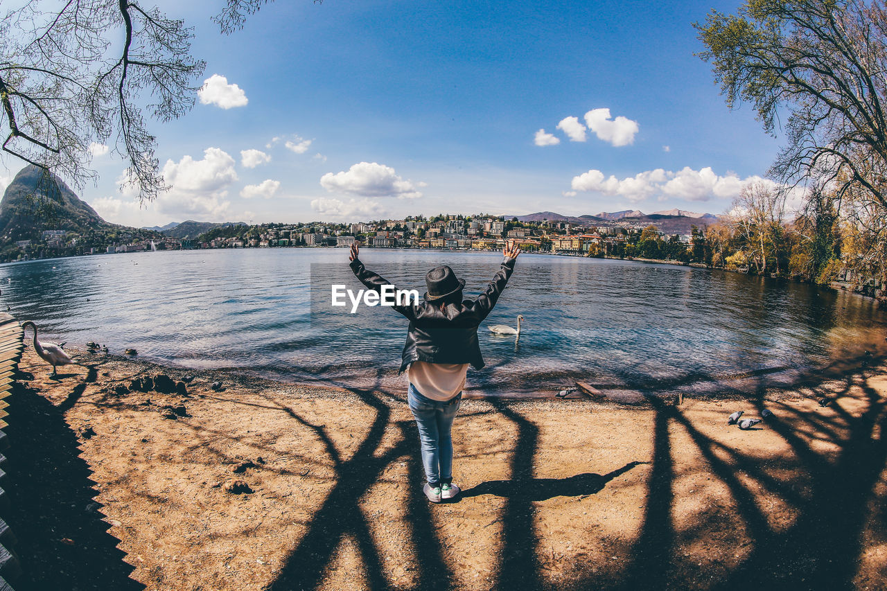 Rear view of cheerful woman standing at lakeshore against sky
