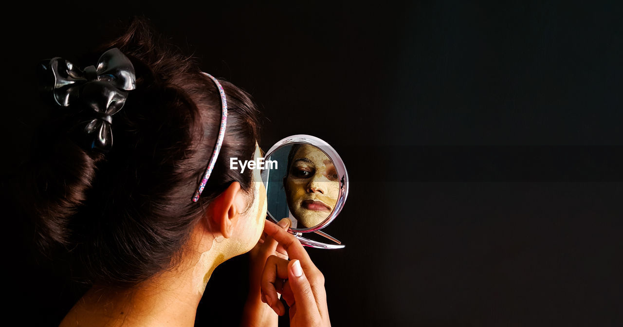 Woman with facial mask holding hand mirror against black background