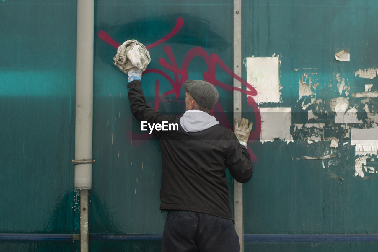 REAR VIEW OF MAN STANDING BY WALL WITH GRAFFITI