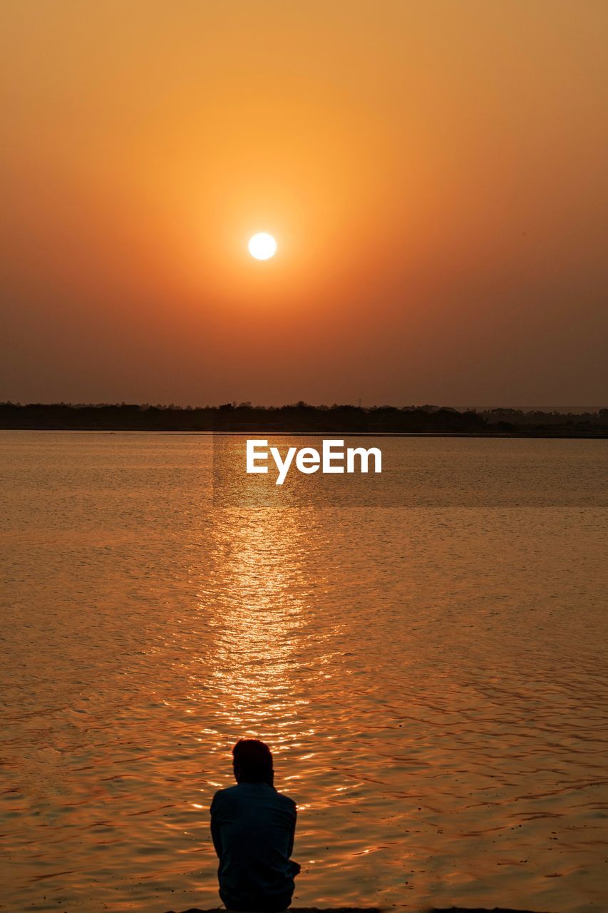 water, sunset, sky, beauty in nature, horizon, tranquility, sea, scenics - nature, nature, orange color, silhouette, tranquil scene, dawn, evening, reflection, sun, one person, relaxation, rear view, sunlight, idyllic, afterglow, vacation, trip, holiday, leisure activity, beach, lifestyles, outdoors, land, sitting, adult, men, non-urban scene, horizon over water, ocean, solitude, environment, travel destinations, landscape, contemplation, travel