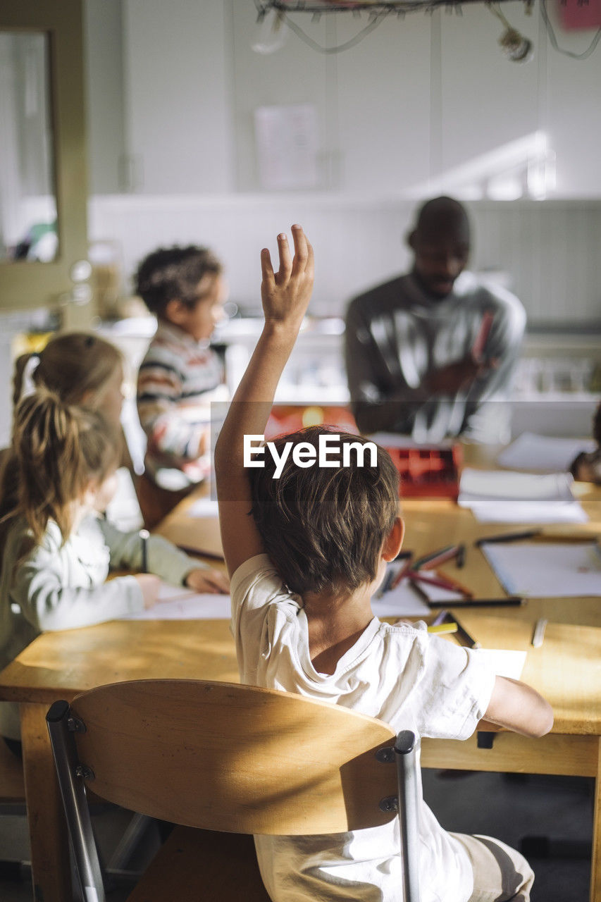 Rear view of boy raising hand while sitting on chair in classroom at kindergarten