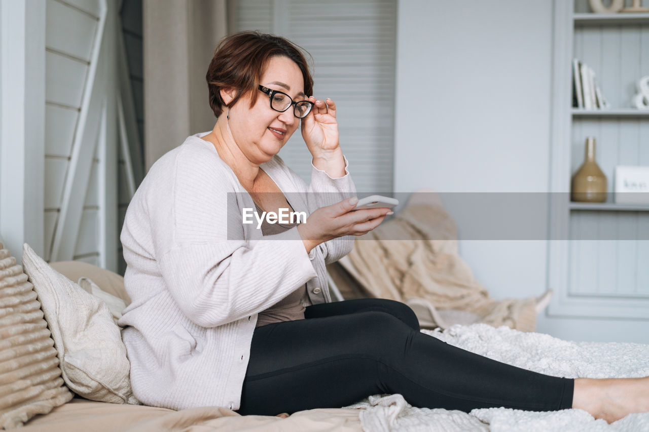 Smiling middle aged plus size woman using mobile phone on bed at home