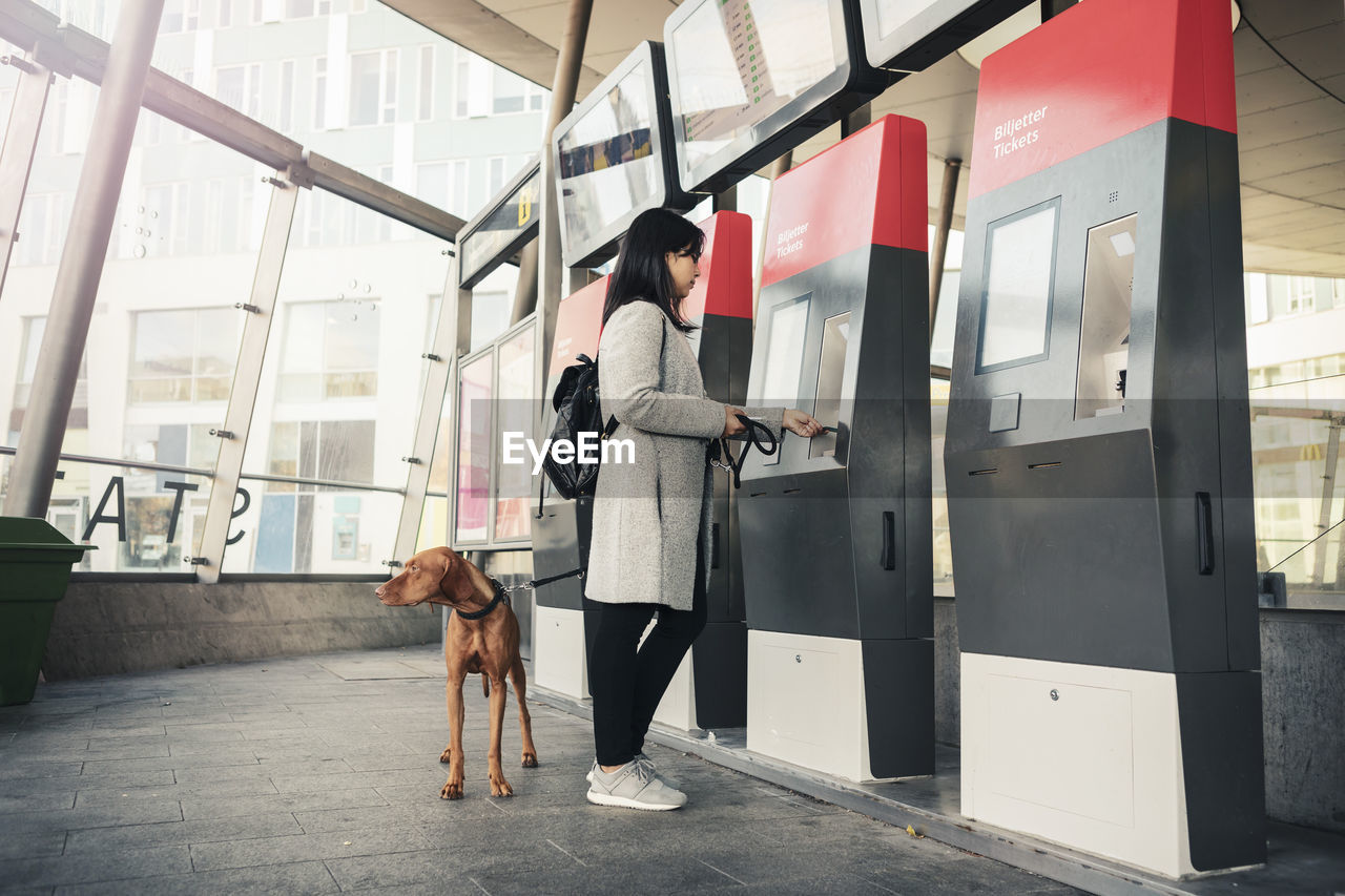 Side view of woman with dog using ticket machine at railroad station