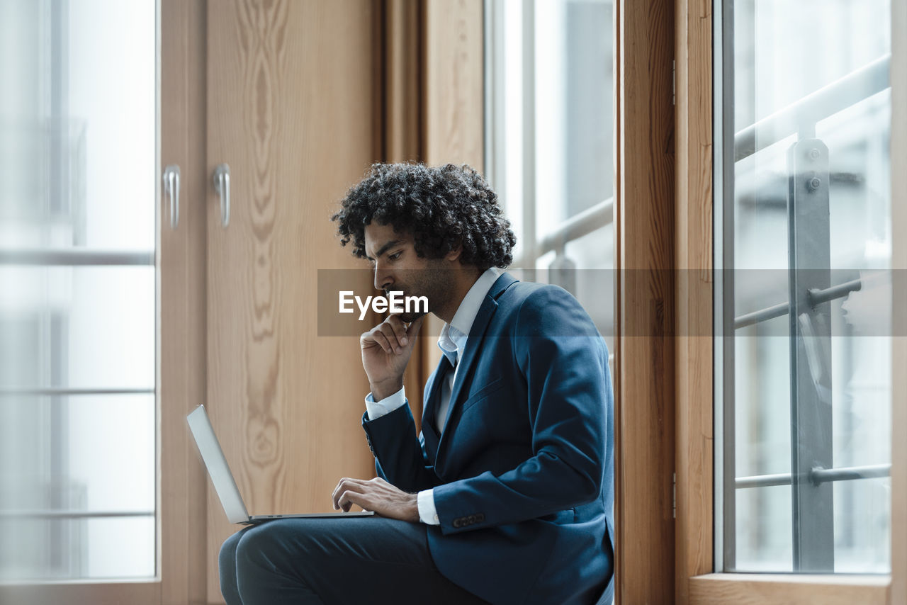 Young male entrepreneur using laptop while sitting against window at workplace