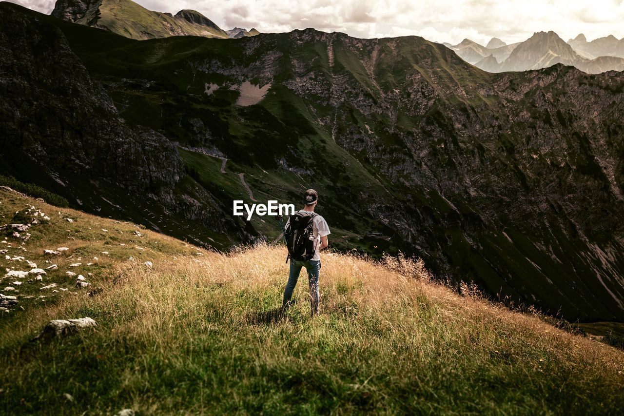 Rear view of man standing on grassy hill against rocky mountains