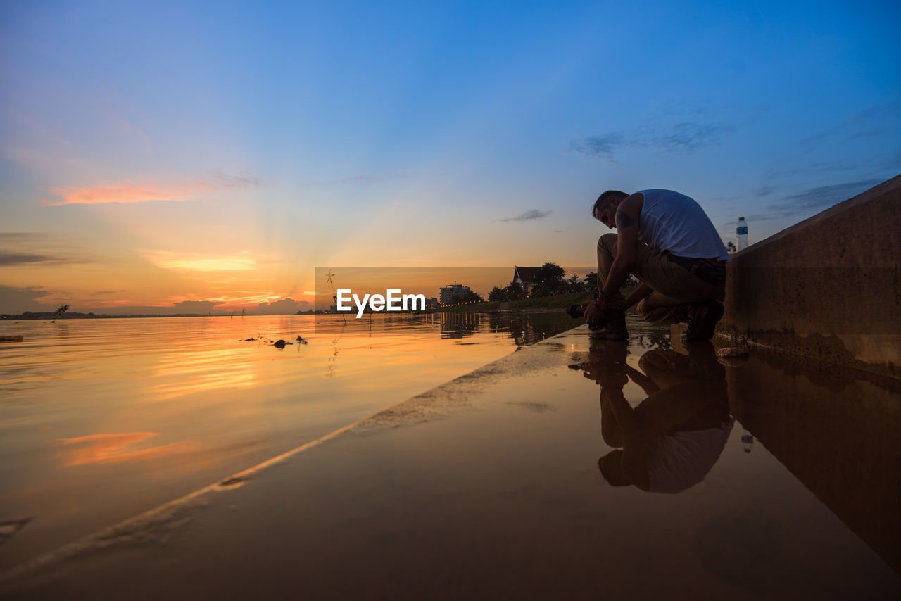 Side view of man crouching by lake against sky during sunset