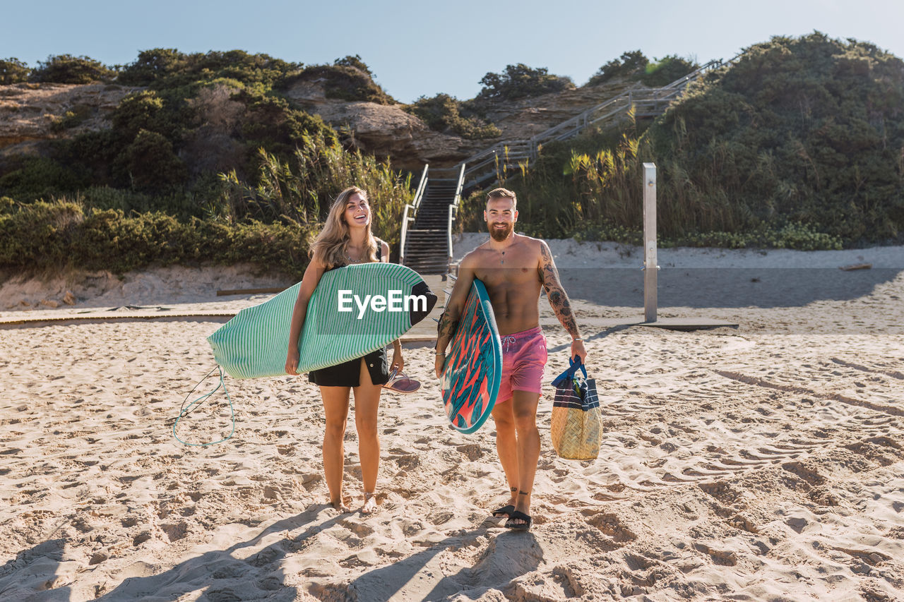 Full body of happy sportive couple with surfboards strolling together on sandy beach before training in the sea