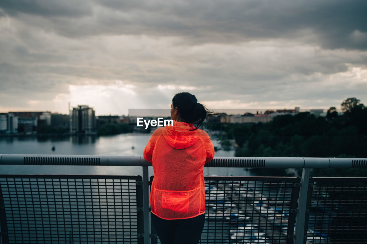 Rear view of female athlete in raincoat looking at city while standing on footbridge over sea during sunset