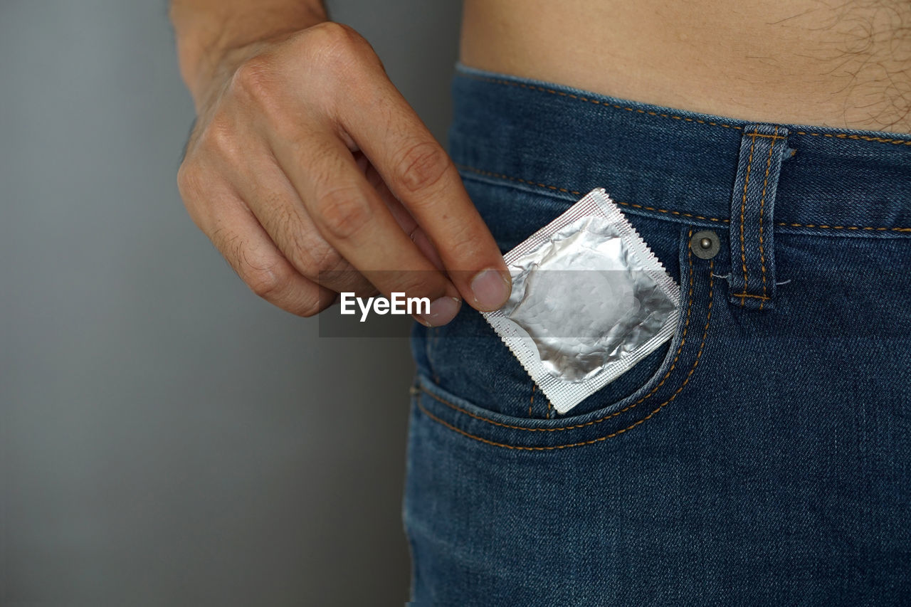 Midsection of man putting condom packet in pocket standing against gray background