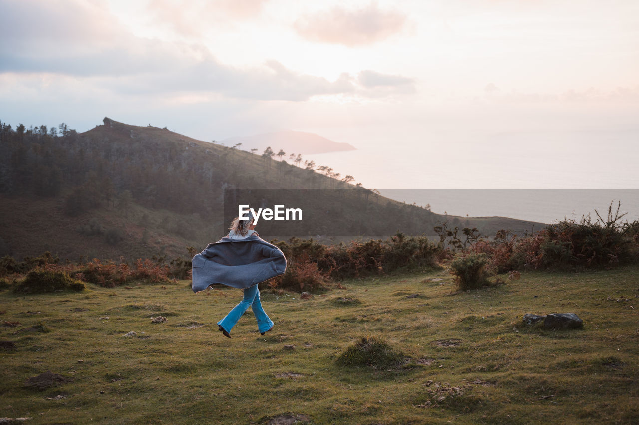 Full body back view of anonymous active female in jacket running on green grassy meadow near mountain ridge in nature