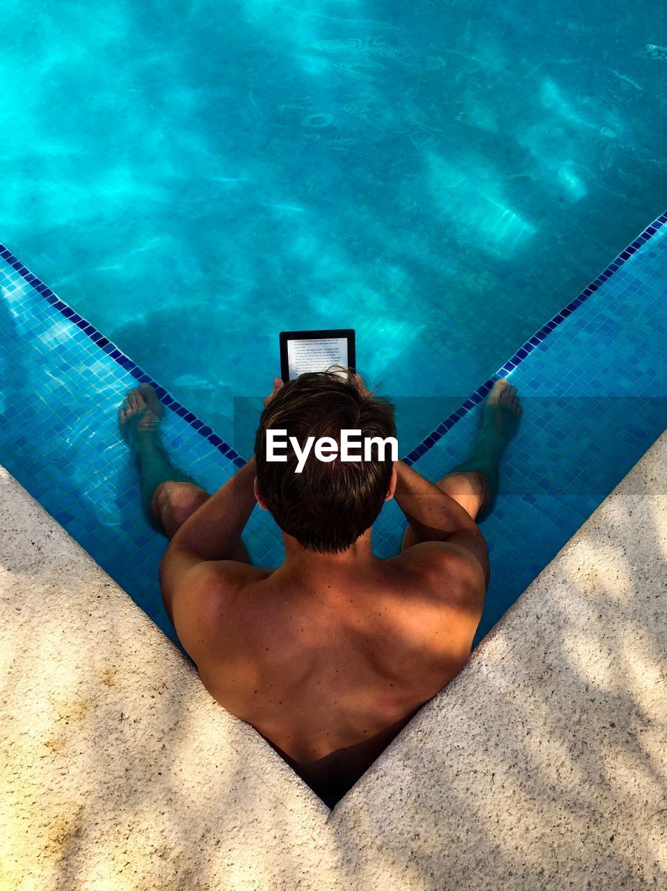 High angle view of shirtless man holding digital tablet while sitting in swimming pool