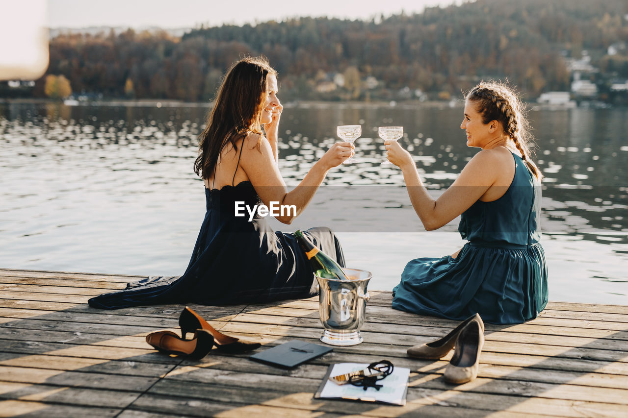 Female event planners toasting champagne while sitting on jetty over lake