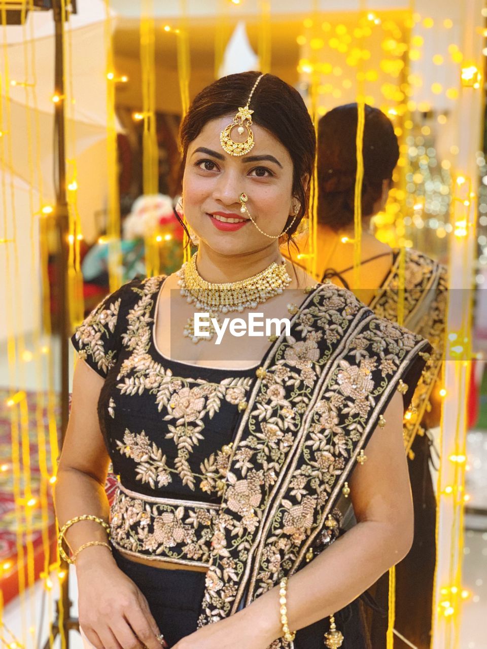 Portrait of smiling beautiful wedding guest wearing traditional clothing and jewelries against illuminated lighting equipment