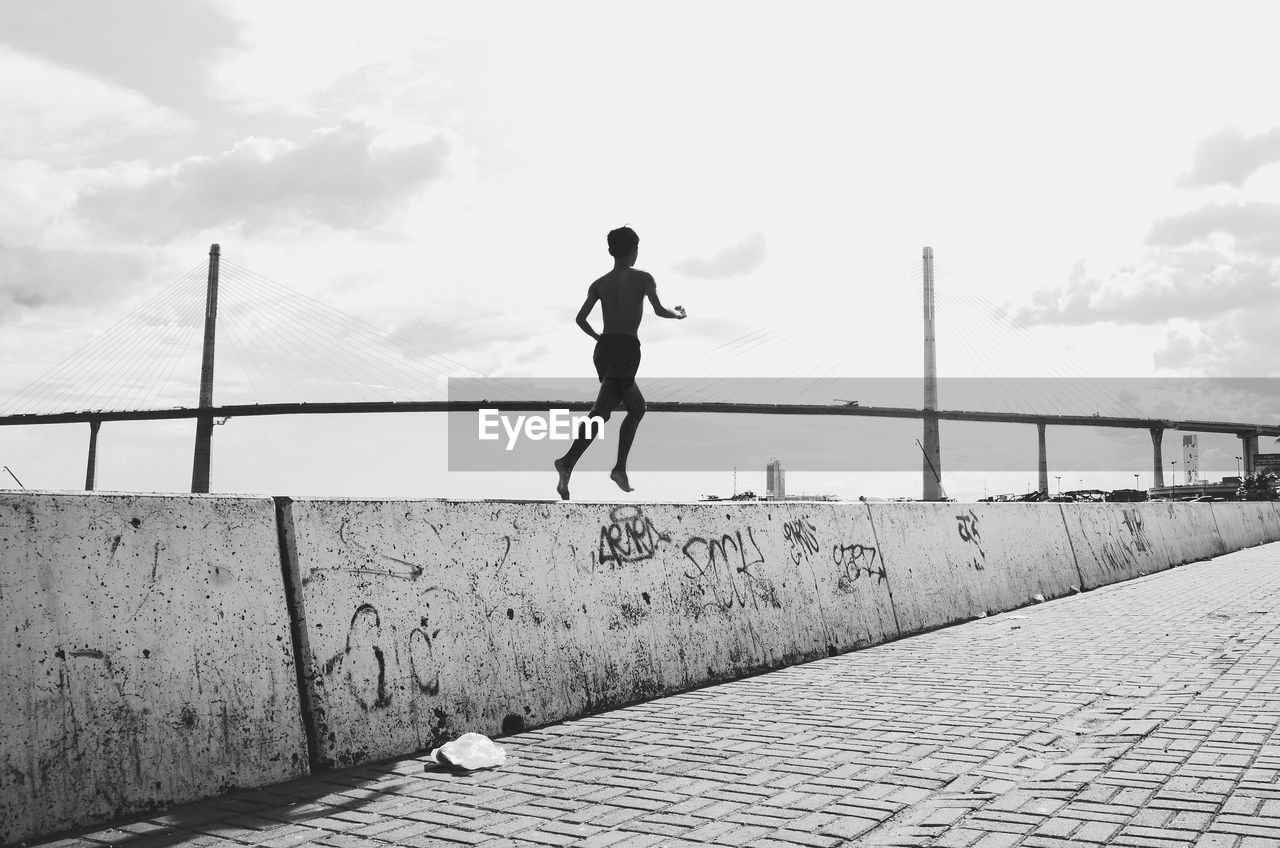 one person, sky, full length, lifestyles, black and white, architecture, cloud, white, leisure activity, sports, monochrome, day, nature, monochrome photography, footpath, men, jogging, city, running, adult, motion, built structure, railing, outdoors, walkway, exercising, black, side view, young adult, bridge, vitality, sunlight, standing, casual clothing, city life, balance, wall, street, water