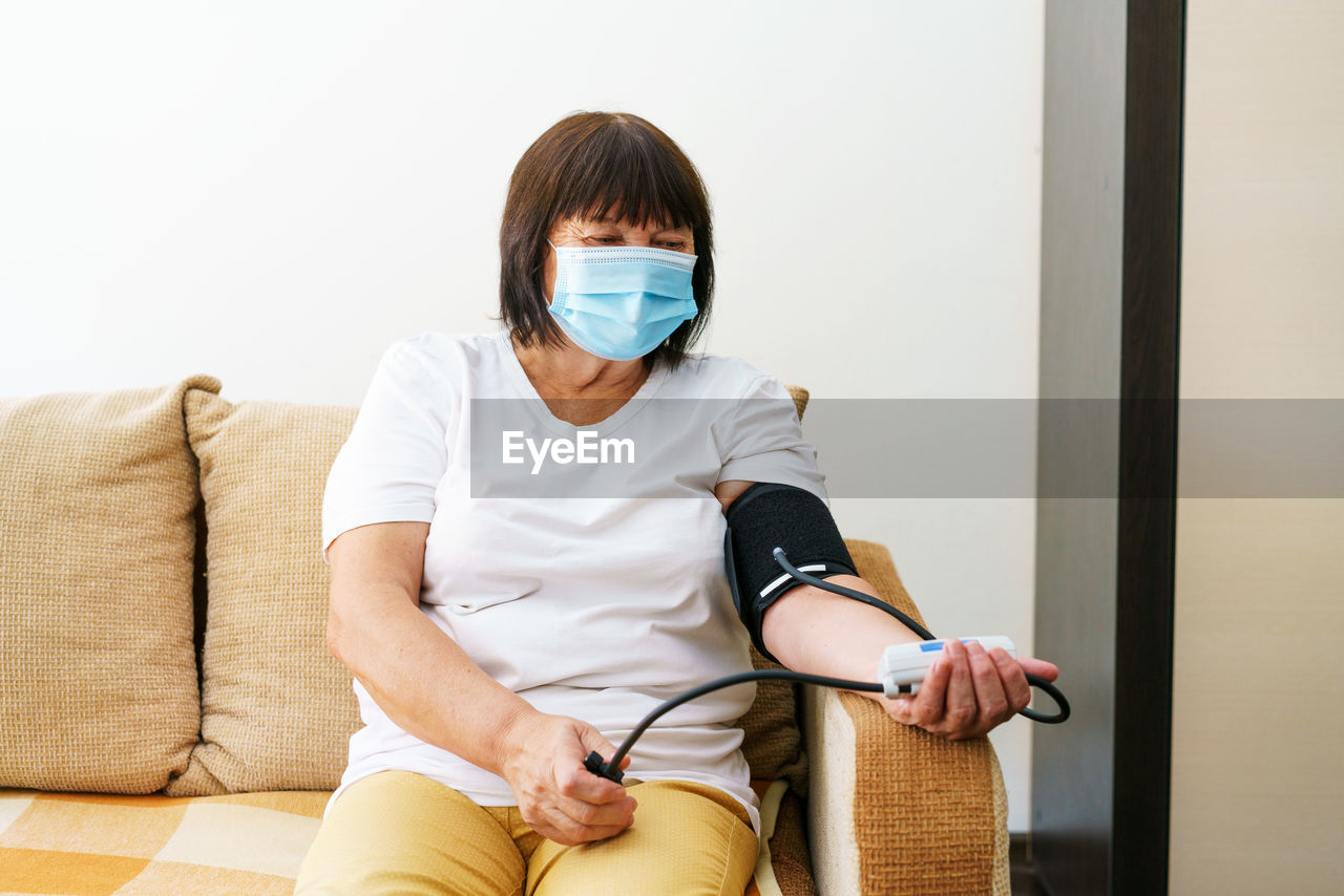 An elderly woman wearing mask measures her blood pressure with an electronic