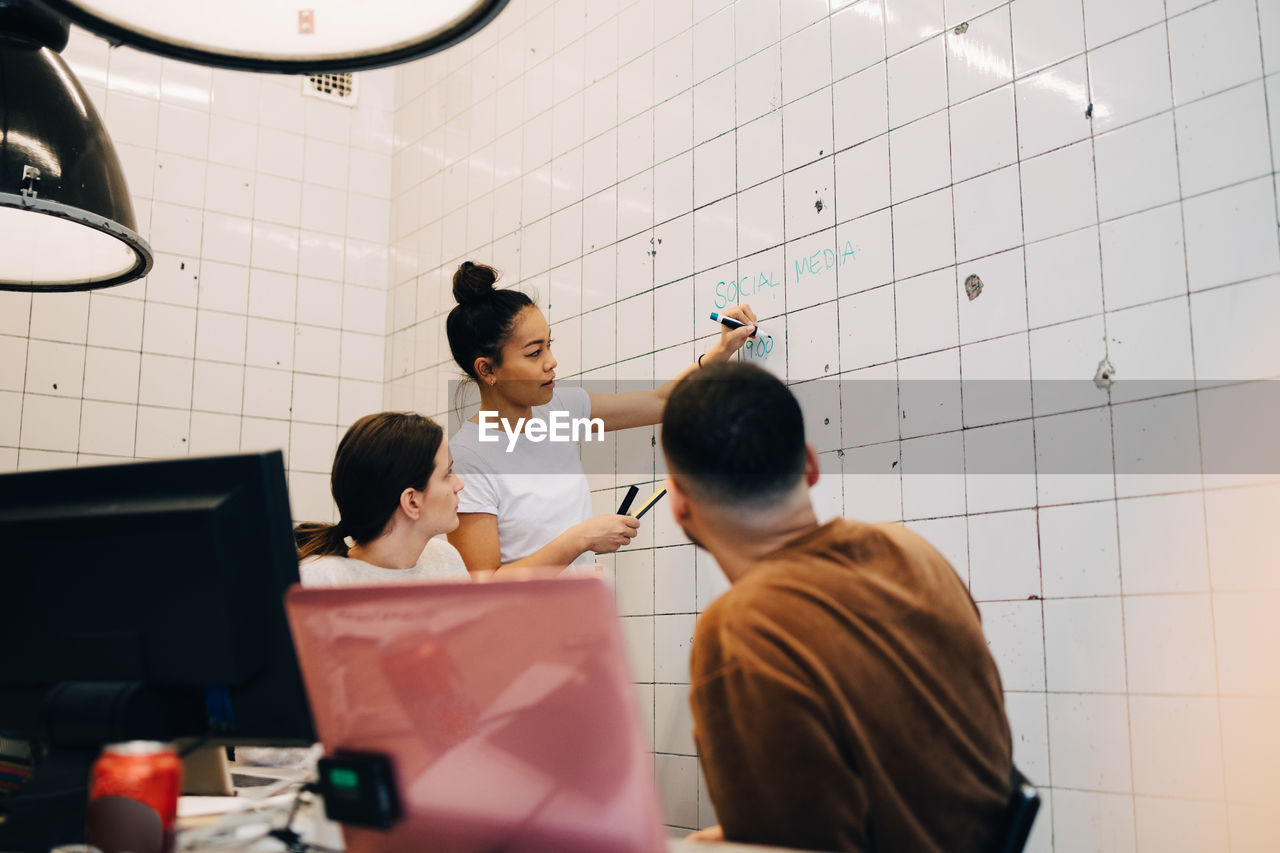 Young businesswoman brainstorming colleagues while writing strategy on tile wall at creative office