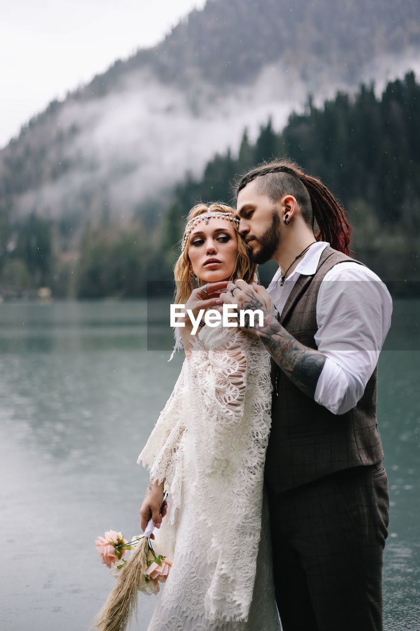 Two happy people in love the bride and groom in wedding outfits embrace by the lake and mountains