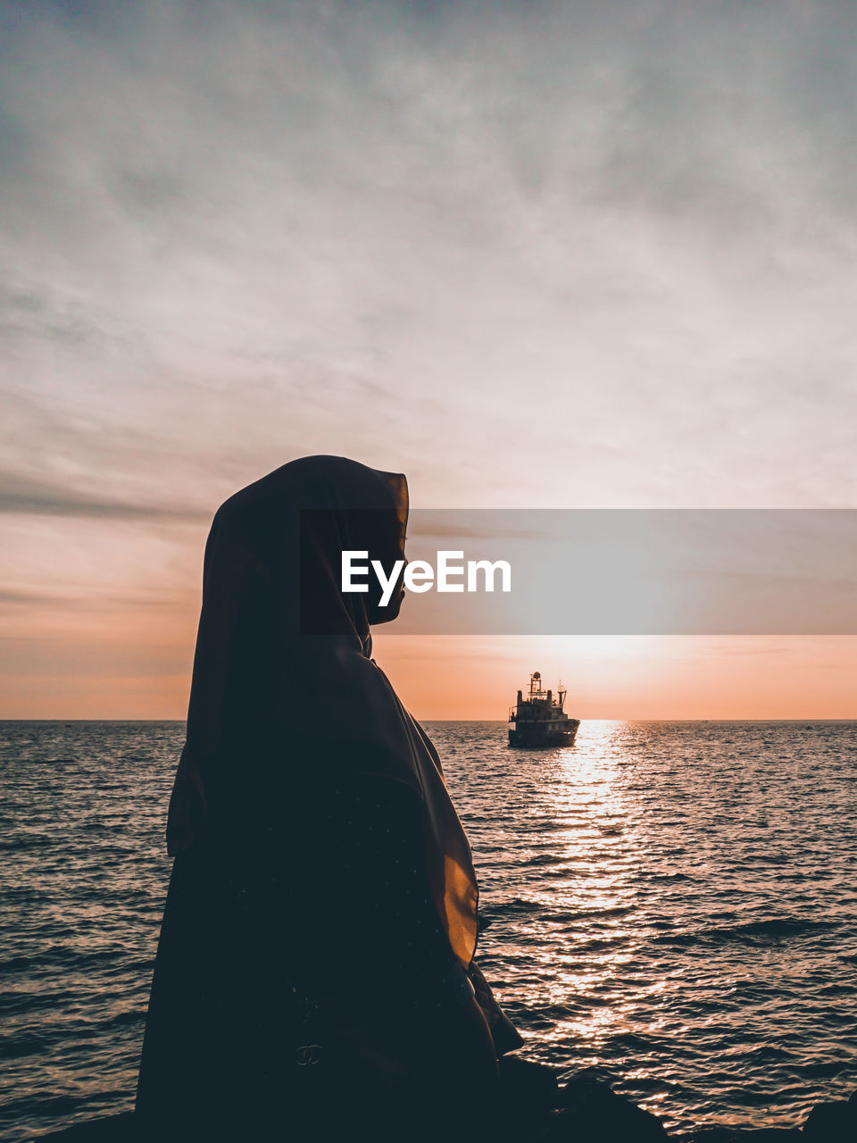 Woman wearing hijab looking away while sitting by sea against sky during sunset