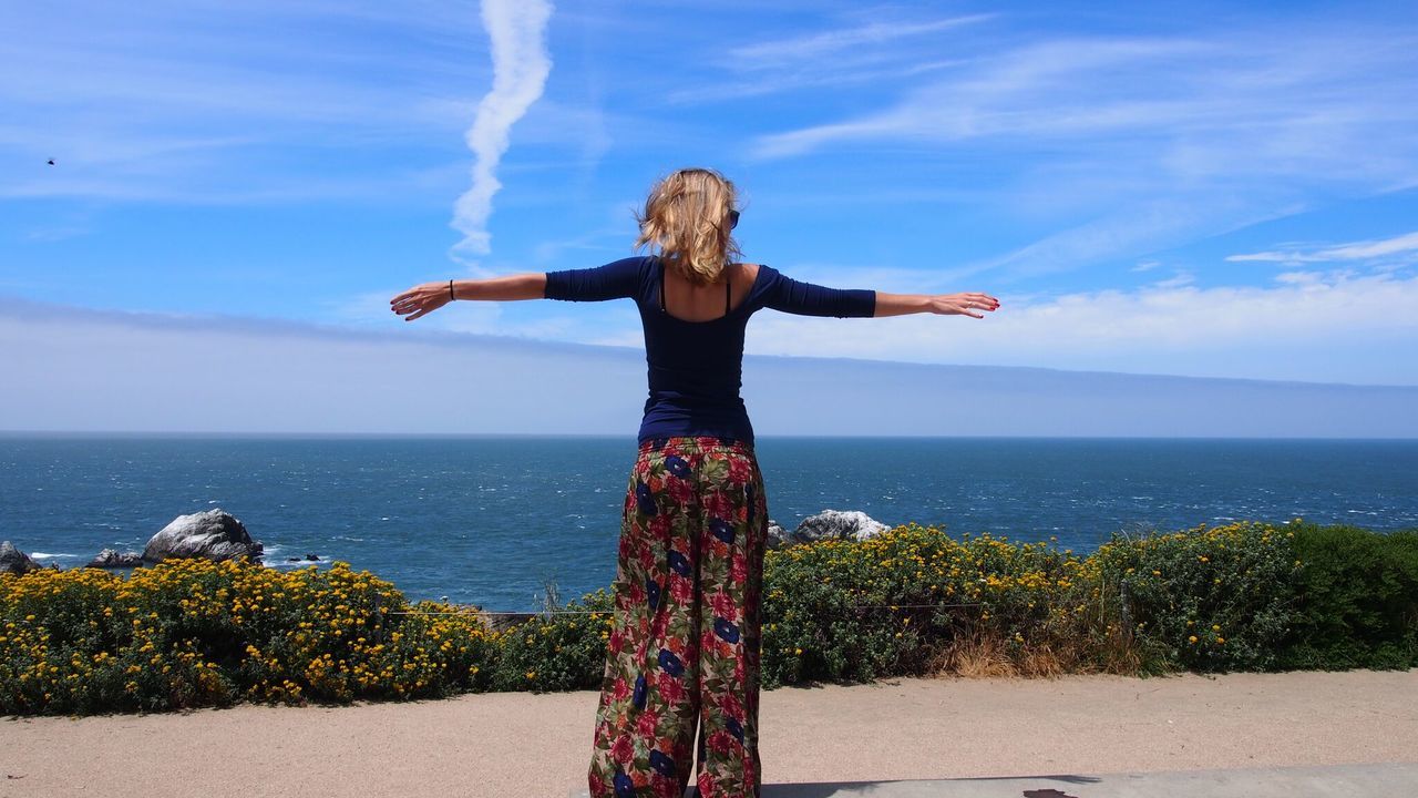 Full length rear view of woman with arms outstretched standing against sea and sky