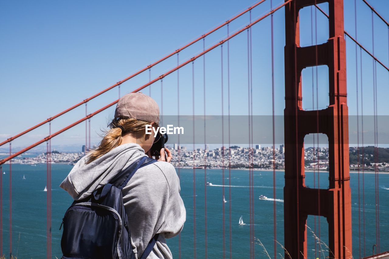 REAR VIEW OF MAN LOOKING AT SUSPENSION BRIDGE AGAINST SKY