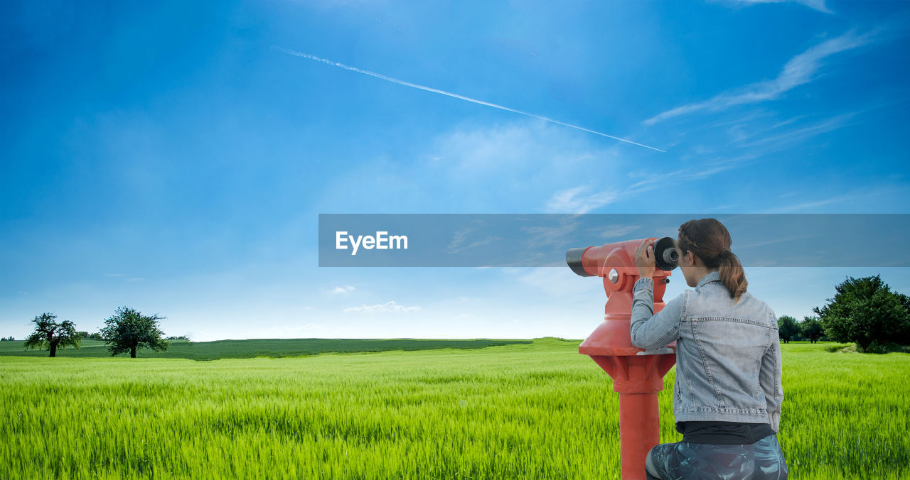 Rear view of woman looking through coin-operated binoculars on field against sky