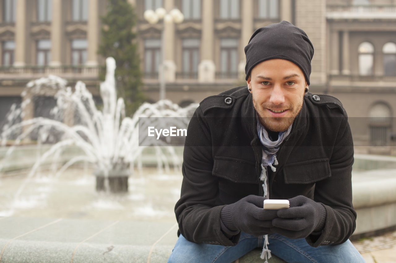 Young man using mobile phone while sitting against water fountain in city