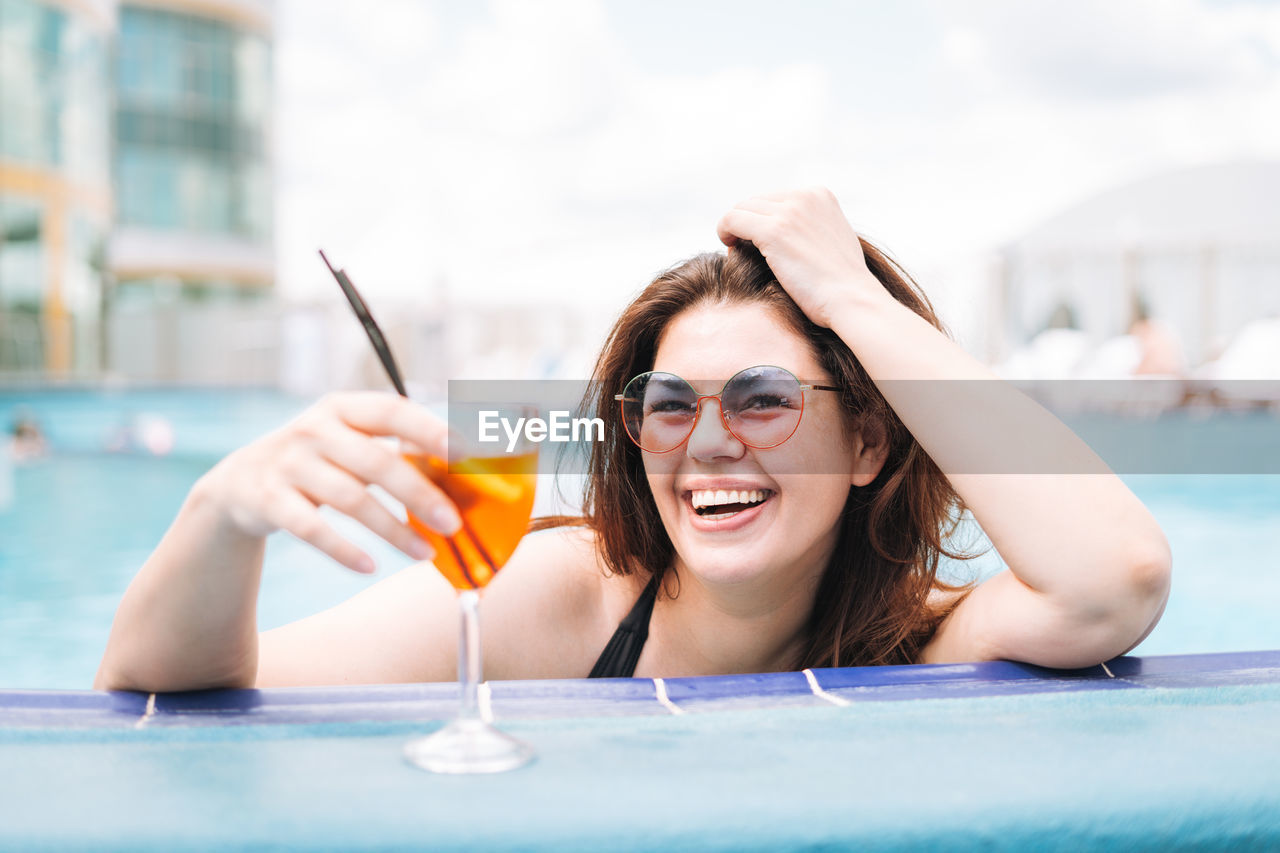 Stylish young woman plus size in black swimsuit and sunglasses with glass of cocktail in pool