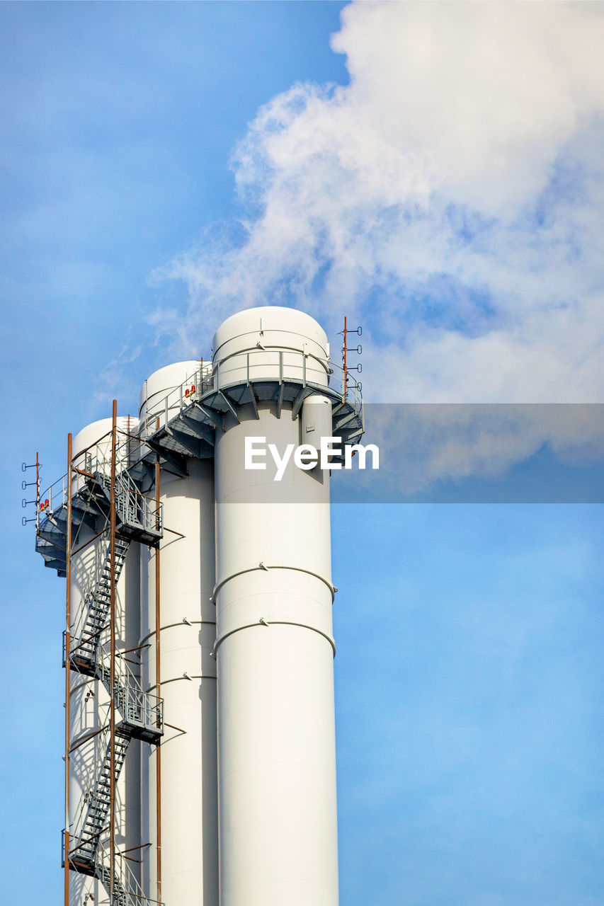 A fragment of pipes of a heating plant emitting white smoke into the blue sky on a bright day. 