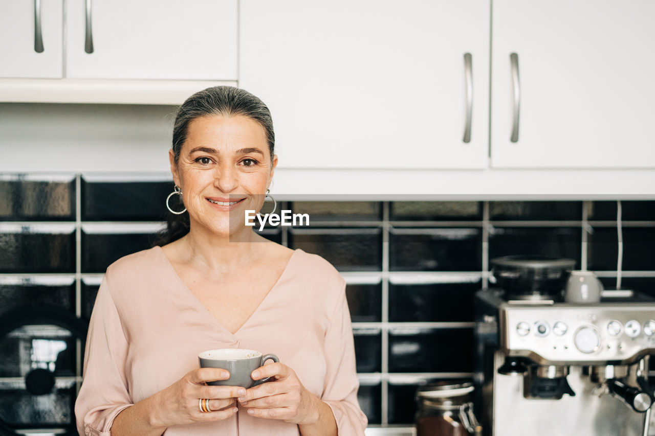 Cheerful middle aged female with mug of hot drink standing at kitchen counter with white cupboards and modern coffee machine at home