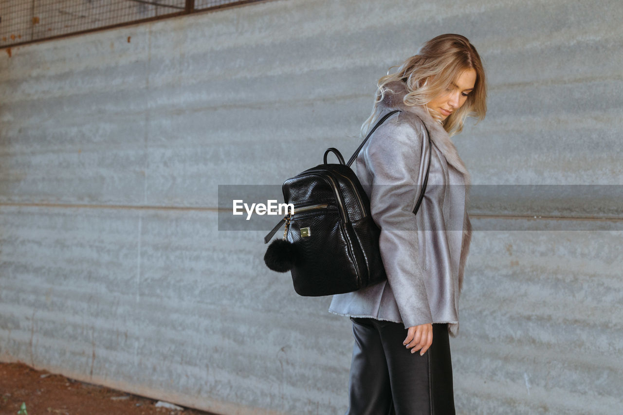 Young woman carrying bag standing against wall