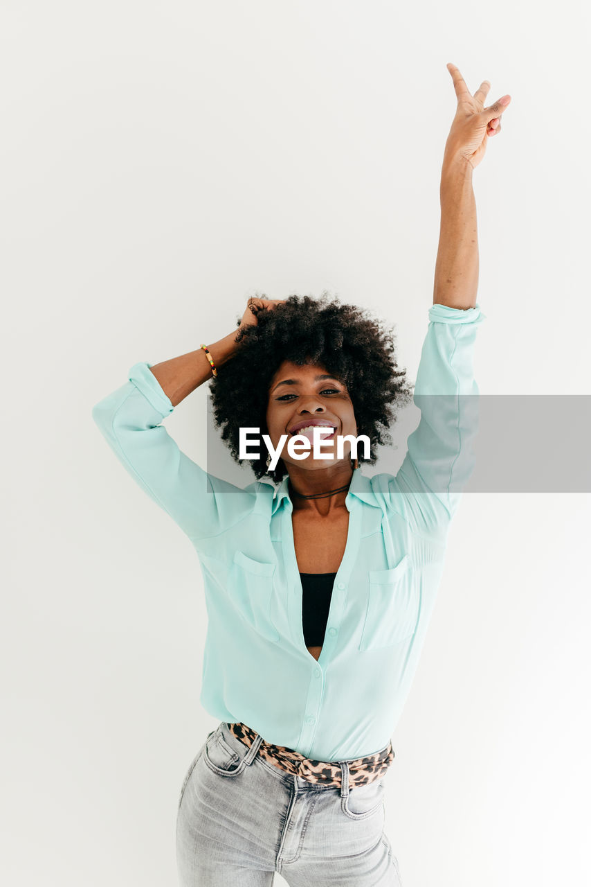 Playful young african american female in trendy outfit having fun and showing tongue and peace sign on white background